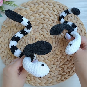 May include: Three crocheted snakes, one black and white, one black, and one white, are displayed on a woven straw mat.