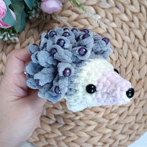 May include: A handmade plush hedgehog toy. The hedgehog has a gray body with purple bead accents, a white and pink face, and black bead eyes. The toy is made of soft, textured material.