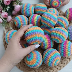 May include: A collection of colorful, crocheted balls in shades of orange, teal, purple, and pink. The balls are arranged on a woven basket, with some held in a person's hand. The handmade spheres are a vibrant display of craftwork.