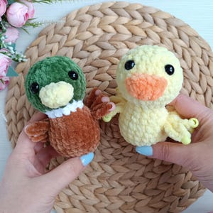 May include: Two handmade crochet duck toys. One duck is yellow with an orange beak, the other is brown, green, and white. Both have black eyes. The toys are on a woven, light brown surface.