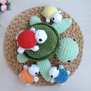 Puede incluir: Una familia de tortugas hechas a mano con ganchillo. La tortuga grande es verde con aletas verde claro. Hay cuatro tortugas más pequeñas en rojo, amarillo, naranja y azul. Las tortugas tienen cabezas blancas y ojos negros. Las tortugas están sobre una estera circular tejida.
