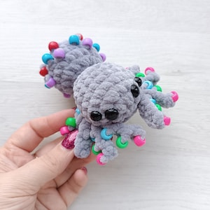 May include: A grey crocheted octopus toy with black eyes and colourful beaded tentacles. The octopus has pink, green, blue, and red beads on its tentacles and head. The plush toy is soft and cuddly.