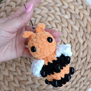 May include: A handmade orange and black crochet bee with large black eyes and white wings. The bee is a plush toy, possibly a keychain or decorative item. The bee is held up by a black string.