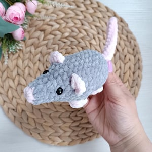 May include: A grey crochet rat with pink detailing on the ears, feet, and tail. The toy rat has black button eyes and a pink nose. The handmade plush is designed to resemble a rodent.