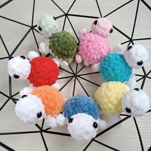 Pu&ograve; includere: Una collezione di otto giocattoli di tartaruga all'uncinetto in vari colori, tra cui rosso, arancione, giallo, verde, blu e rosa. Le tartarughe sono disposte in cerchio su uno sfondo di rete metallica nera.