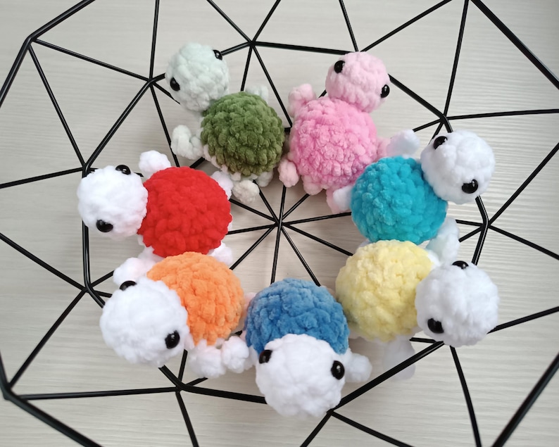 Pu&ograve; includere: Una collezione di otto giocattoli di tartaruga all'uncinetto in vari colori, tra cui rosso, arancione, giallo, verde, blu e rosa. Le tartarughe sono disposte in cerchio su uno sfondo di rete metallica nera.