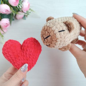 May include: A handmade, plush, light brown and tan capybara toy with closed eyes and a black stitched mouth. A red heart-shaped plush is held nearby. The capybara is approximately 4 inches long.