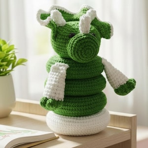 May include: A handmade crochet toy in the shape of a green and white dragon. The dragon has a round head, a stacked body, and white accents on its horns, scarf, and feet. The toy is sitting on a wooden surface.