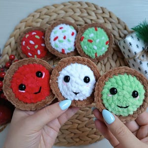 May include: Five crocheted plush cookies with different toppings, including red and white sprinkles, green frosting, and red frosting. Each cookie has a smiling face with black eyes.