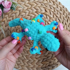 May include: A turquoise crocheted dinosaur toy with colorful bead accents. The handmade plush toy features a textured surface and is decorated with blue, orange, pink, and green beads. The dinosaur has a playful design, perfect for children.