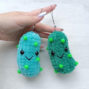 May include: Two plush pickle charms made of soft, textured yarn in light and dark green shades. Each pickle features black plastic safety eyes, a stitched black smile, and small, bright green plastic beads attached to the surface. Each charm includes a black cord loop for hanging.