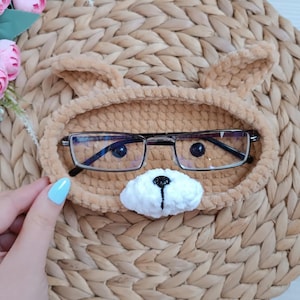 May include: A crocheted brown and white bear-shaped glasses holder with a white nose and black eyes. The holder is sitting on a woven brown surface.