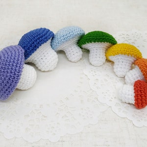 K&ouml;nnte beinhalten: Ein Set aus sieben geh&auml;kelten Pilzen in Regenbogenfarben. Die Pilze sind in einem Halbkreis auf einer wei&szlig;en Spitze angeordnet.