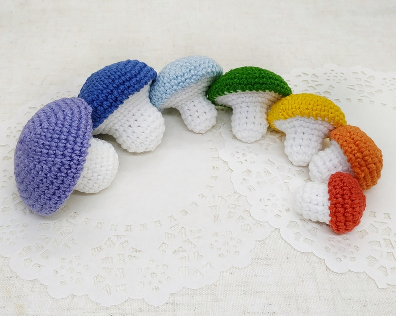 K&ouml;nnte beinhalten: Ein Set aus sieben geh&auml;kelten Pilzen in Regenbogenfarben. Die Pilze sind in einem Halbkreis auf einer wei&szlig;en Spitze angeordnet.