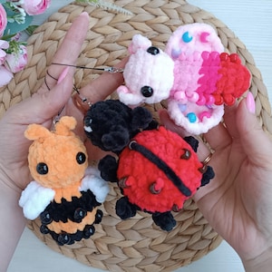 Könnte beinhalten: Eine Sammlung handgefertigter Häkel-Schlüsselanhänger in Form einer Biene, eines Marienkäfers, eines Schmetterlings und zweier kleiner Kreaturen. Die Biene ist orange und schwarz, der Marienkäfer ist rot und schwarz, und der Schmetterling ist rosa und rot. Jeder Schlüsselanhänger hat schwarze Perlenaugen.