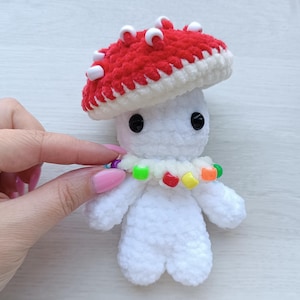May include: A handmade crochet mushroom doll. The mushroom has a red cap with white accents, a white body, black eyes, and a colourful beaded necklace. The doll is held by a person.