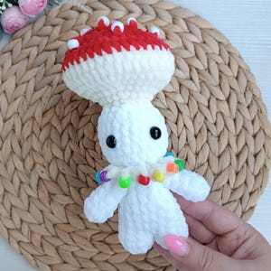 Könnte beinhalten: Handgemachte Häkelpilzpuppe mit rotem und weißem Hut, weißem Stiel und schwarzen Augen. Die Puppe hat kleine Arme und eine bunte Perlenkette. Der Pilz ist aus weichem, strukturiertem Garn gefertigt.