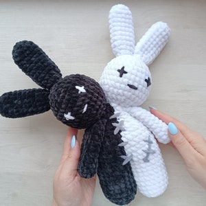 May include: Two crocheted bunny plushies, one black and one white, with stitched-on eyes and a smiling expression. The plushies are joined together at the waist.