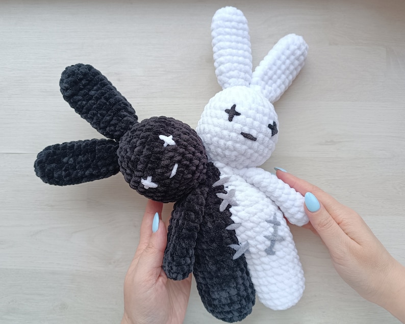 May include: Two crocheted bunny plushies, one black and one white, with stitched-on eyes and a smiling expression. The plushies are joined together at the waist.