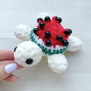 Puede incluir: Un juguete de tortuga de ganchillo hecho a mano con forma de rodaja de sandía. La tortuga tiene una parte superior roja con semillas de cuentas negras, ribetes blancos y verdes y extremidades blancas y esponjosas. El juguete está diseñado para parecerse a una sandía.
