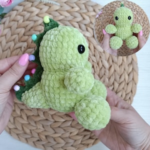 May include: A light green, plush, crocheted dinosaur toy with a textured surface. The dinosaur has a black button eye and a green spine with colorful bead accents. A smaller version of the dinosaur is also shown.