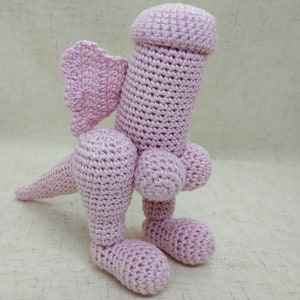 Könnte beinhalten: Ein rosa gehäkeltes Drachenspielzeug mit einem langen Schwanz und einem Flügel auf einer Seite. Der Drache hat einen langen Hals und einen runden Kopf.