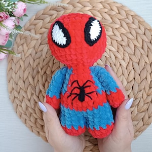 May include: A red and blue crocheted plush toy of Spider-Man. The toy has a black spider symbol on its chest and white eyes.