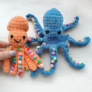 May include: Two handmade crochet octopus toys. One is orange, the other is blue. Both have black eyes and beaded tentacles in various colors. The orange octopus is held by a hand with a glittery nail. The toys are on a light surface.