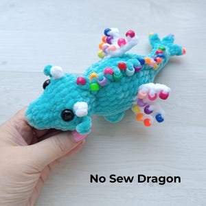 Könnte beinhalten: Ein türkisfarbenes gehäkeltes Drachenspielzeug mit schwarzen Augen und weißen Akzenten. Der Drache ist mit bunten Perlen auf dem Rücken und den Flügeln verziert. Der Text "No Sew Dragon" ist unten im Bild zu sehen.