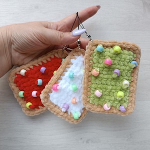 May include: Three crocheted, rectangular charms resembling frosted pastries. One is red, one is white, and one is green, each with colorful bead sprinkles and a tan border. They are attached to a black string.