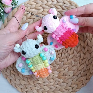 May include: Two handmade crochet butterfly charms. One is light blue with rainbow accents and the other is pink with red and orange accents. Both have black eyes and are embellished with colourful beads. Each charm has a black string attached.