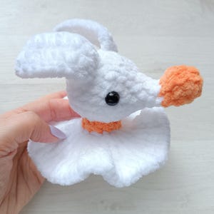 Pode incluir: Um brinquedo de ganso de crochê branco feito à mão com uma textura fofa. O ganso tem um bico laranja brilhante e uma gola laranja combinando. Possui um olho preto e orelhas grandes e caídas.