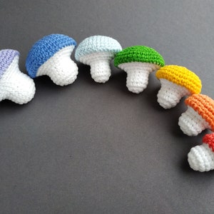 K&ouml;nnte beinhalten: Ein Regenbogen aus geh&auml;kelten Pilzen in verschiedenen Gr&ouml;&szlig;en. Die Pilze sind aus wei&szlig;em Garn mit unterschiedlichfarbigen Oberseiten, darunter lila, blau, hellblau, gr&uuml;n, gelb, orange und rot.