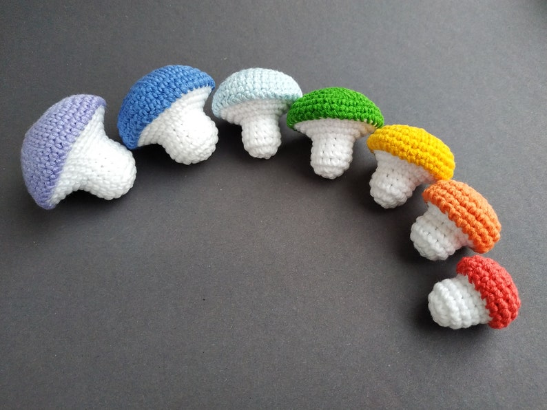 K&ouml;nnte beinhalten: Ein Regenbogen aus geh&auml;kelten Pilzen in verschiedenen Gr&ouml;&szlig;en. Die Pilze sind aus wei&szlig;em Garn mit unterschiedlichfarbigen Oberseiten, darunter lila, blau, hellblau, gr&uuml;n, gelb, orange und rot.