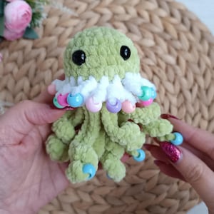 May include: A handmade, green crocheted octopus plush toy with black eyes and a white frill adorned with colorful beads. The tentacles have blue beads at the tips. A whimsical, soft toy.