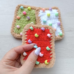 May include: Crocheted square-shaped items resembling pastries. One is red with colorful bead sprinkles, held by a hand. Two others are green and white, also with sprinkles. Each has a tan border, suggesting a food-themed craft.