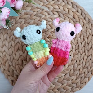 Könnte beinhalten: Zwei handgefertigte Häkeltiere im Bild. Eines ist hellblau mit einem Regenbogenbauch und türkisfarbenen Perlen. Das andere ist rosa mit einem roten Bauch. Beide haben schwarze Augen und kleine Hörner. Die Kreaturen werden in einer Hand gehalten.