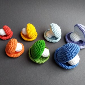 K&ouml;nnte beinhalten: Ein Set aus sieben geh&auml;kelten Muscheln in verschiedenen Farben, darunter Rot, Orange, Gelb, Gr&uuml;n, Blau und Lila. Jede Muschel enth&auml;lt eine wei&szlig;e Perle.