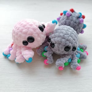 May include: Two crocheted octopus toys. One is pink with black eyes and blue and pink accents. The other is gray with black eyes and green, pink, and blue accents. The toys are soft and plush.