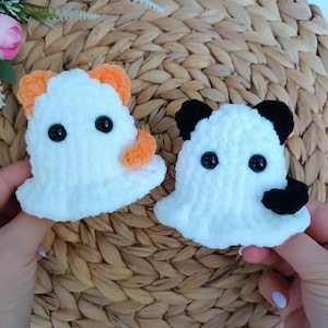 May include: Two crocheted ghost figures made of white yarn. One ghost features orange ears and an orange arm, while the other has black ears and a black arm. Both figures have round, black plastic eyes and a soft, textured, plush appearance.