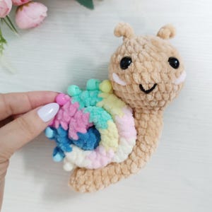 May include: A crocheted snail toy with a tan body, black eyes, and a smiling face. The snail's shell is a spiral of pastel colours including pink, blue, and yellow. The toy is made of soft, textured yarn.