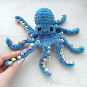 Könnte beinhalten: Handgefertigter blauer Häkel-Oktopus mit schwarzen Sicherheitsaugen und bunten Perlen an den Tentakeln. Das weiche Spielzeug ist strukturiert und eignet sich für ein Kinderzimmer oder als Dekoration. Ein einzigartiges, handgefertigtes Produkt.