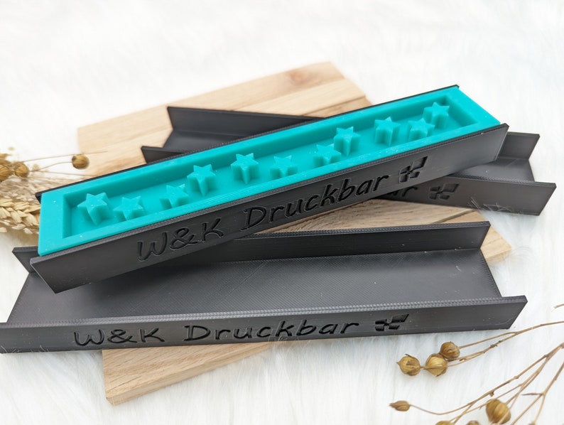 May include: Two black plastic moulds with the text "W&K Druckbar" on them. The top mould has a turquoise blue insert with a row of five star shapes.