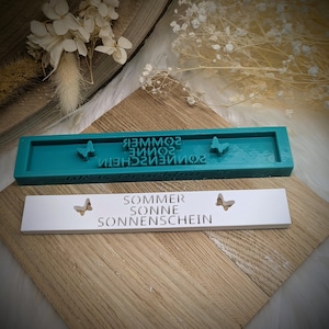 Könnte beinhalten: Eine türkisfarbene rechteckige Form mit dem Text "SOMMER SONNENSCHEIN" und Schmetterlingsausschnitten, zusammen mit einem weißen rechteckigen Schild mit demselben Text und Schmetterlingsausschnitten. Die Form befindet sich auf einer Holzoberfläche.