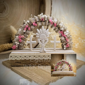 Könnte beinhalten: Weißer Holzbogen, verziert mit rosa und weißen Blumen, zwei weißen Kreuzen und einem weißen Kelch mit Sonnenstrahlen-Design. Der Bogen steht auf einem Holzsockel mit einer Spitzenverzierung.