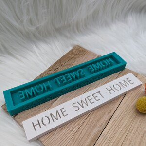 May include: Two rectangular silicone moulds for crafting. The top mould is teal blue and says "Home Sweet Home" in white lettering. The bottom mould is white and says "Home Sweet Home" in black lettering.