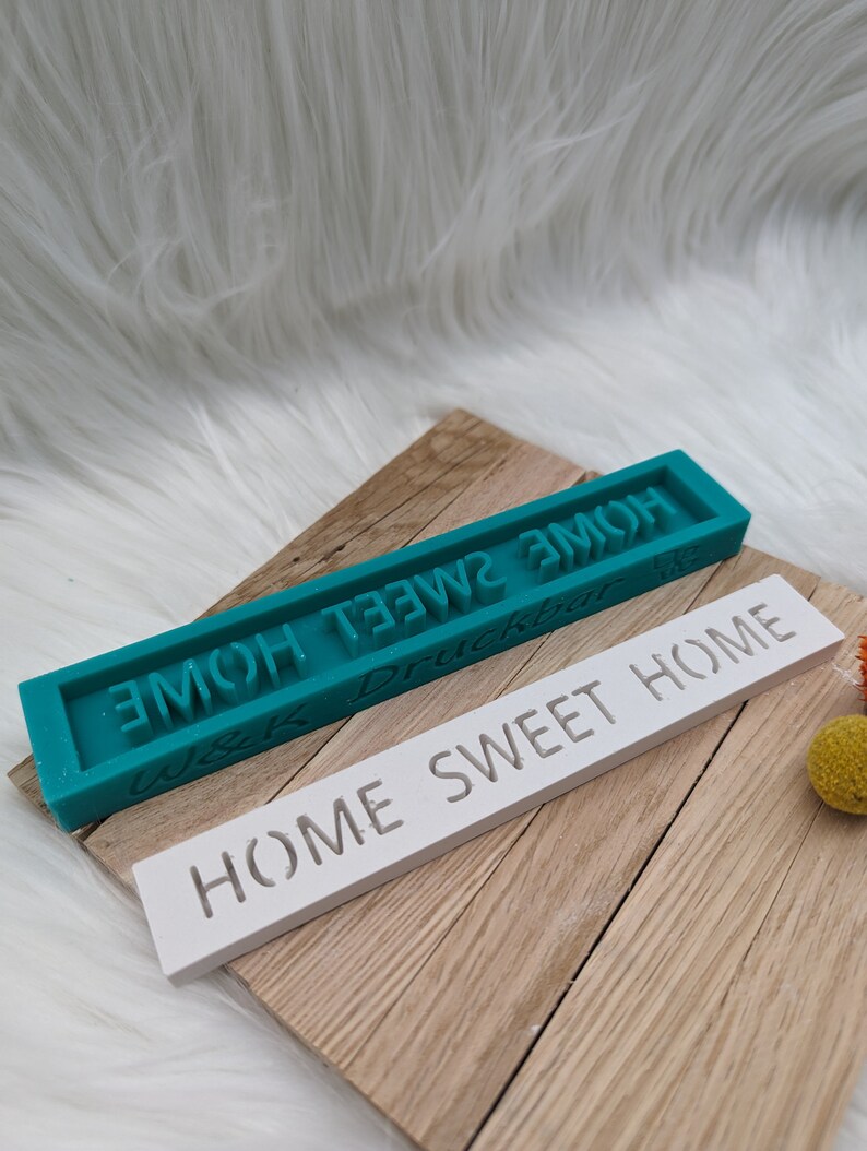 May include: Two rectangular silicone moulds for crafting. The top mould is teal blue and says "Home Sweet Home" in white lettering. The bottom mould is white and says "Home Sweet Home" in black lettering.