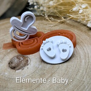 Könnte beinhalten: Ein weißer Schnuller und ein Baby-Fußabdruck-Stempel mit Herz-Ausschnitten werden gezeigt. Die Artikel befinden sich auf einem braunen Hintergrund. Der Text "Elemente - Baby -" ist unten im Bild zu sehen.