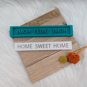 May include: A teal blue silicone mould with the text "HOME SWEET HOME" in white lettering. The mould is sitting on a wooden surface with a white stencil that reads "HOME SWEET HOME" on top of it.
