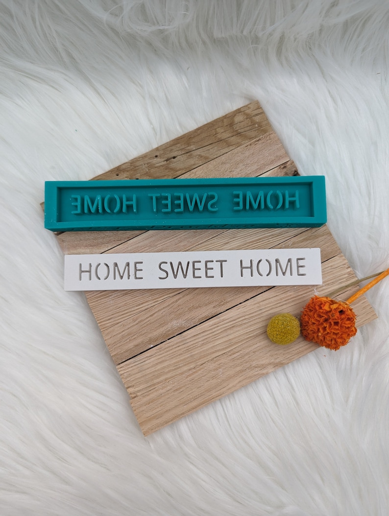 May include: A teal blue silicone mould with the text "HOME SWEET HOME" in white lettering. The mould is sitting on a wooden surface with a white stencil that reads "HOME SWEET HOME" on top of it.